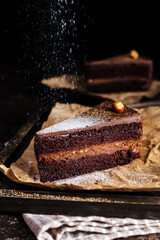 A beautiful piece of chocolate cake with cream and ganache on a wooden board. Powdered sugar falls beautifully on a cake with nuts