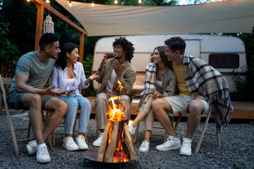Happy multinational friends sitting near campfire, talking to each other, enjoying warm summer...