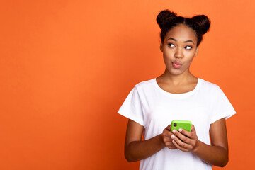 Photo of young pretty african woman use smartphone wondered look empty space thoughtful isolated over orange color background