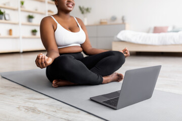 Plus size black woman having online meditation or yoga class, sitting in lotus pose next to laptop...