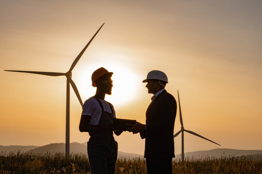 Silhouette of african american engineer and indian inspector using digital tablet during meeting outdoors. Two partners standing on field with wind turbines during amazing summer sunset.