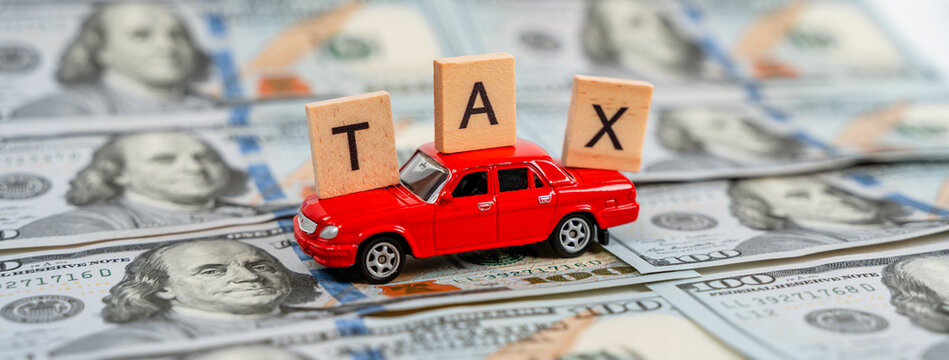 A toy car carries the wooden letters "tax" standing on dollar bills. The concept of paying taxes for a car, buying a new car