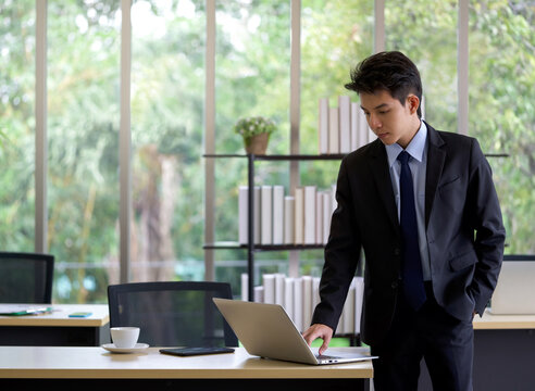 Young Asian Businessman In Black Suit Use Laptop Computer To Check Email Before Go Out To Lunch Break.