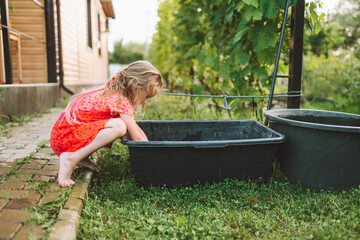 Little barefooted blond girl in red dress sit on haunches putting hands into green plastic...