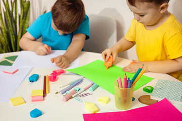 Happy kids with yellow and blue shirts doing arts and crafts together at their desk. Children draw on colored paper. Boys play at home.