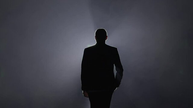 Silhouette of a man in a suit leaves the stage 4K. Mystical atmosphere, smoke, spotlight. Person in the dark