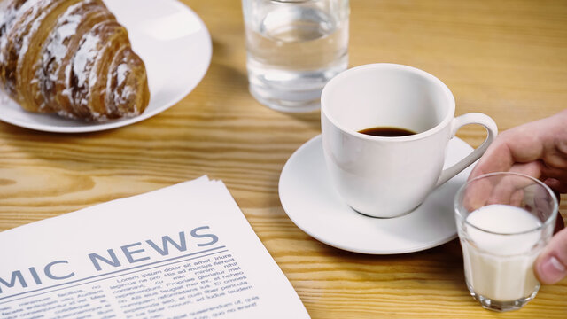 cropped view of man taking glass of milk near cup of coffee, newspaper and croissant.