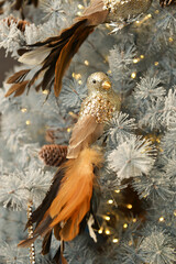 A golden Christmas tree toy on a clothespin in the shape of a bird with a tail of red and brown feathers sits on an artificial snow-covered white Christmas tree with burning lights of a garland.