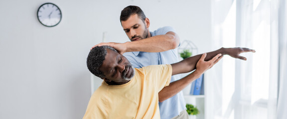 physiotherapist stretching arm of african american man while examining him in rehab center, banner