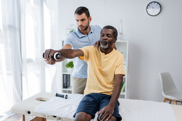 physical therapist supporting african american man working out with dumbbell while sitting on...