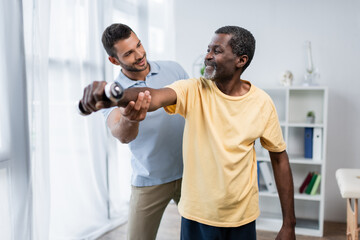 young trainer assisting happy african american man training with dumbbell in rehabilitation center - obrazy, fototapety, plakaty