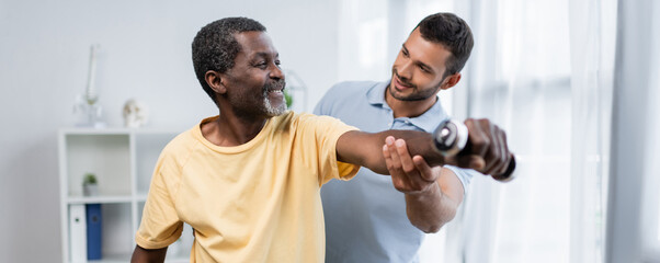 young physiotherapist assisting smiling african american patient exercising with dumbbell, banner