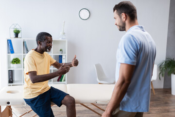 Obraz premium mature african american man showing thumbs up while talking to rehabilitologist in clinic