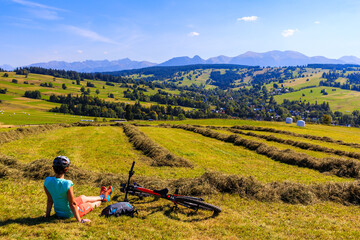 Young woman sitting on meadow with bike and looking at beautiful panorama of Tatra Mountains, Poland - obrazy, fototapety, plakaty