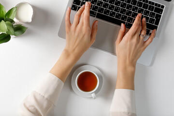 hands of a woman typing on a laptop on a white table with a cup of tea. Top view.