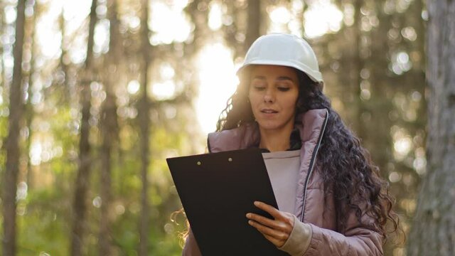 Millennial experienced Female technician with clipboard taking measures for reforestation of woodlands. Young indian Forestry engineer in hardhat in park. Supervising wildlife sanctuary checking trees - Powered by Adobe