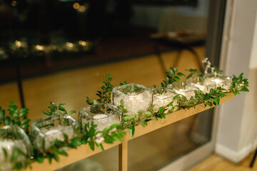 A row of candles in transparent glass holders with greenery lined up on the table Wedding reception decoration Cropped photo Side view Close up