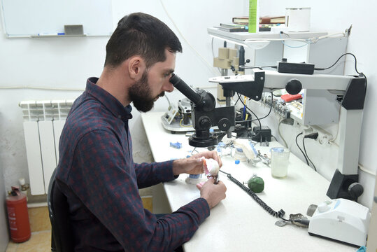 A Young Man Looks Through A Microscope Examining Repairs Dentures Or A Jaw In The Workshop Of A Dental Technician