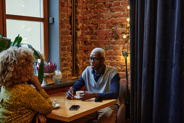 African young businessman in eyeglasses drinking coffee at the table together with woman during their meeting in coffee shop