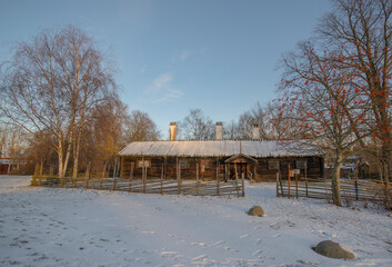 Old log stable and houses with birch-bark roofs in a snowy park a sunny winter day afternoon in...