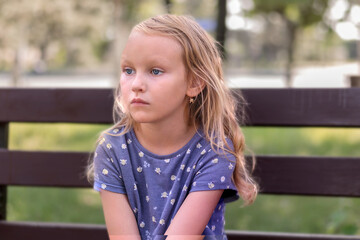 little girl, blonde, sits with a displeased look, angry, on a bench in the park