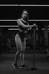 Fototapeta na wymiar A full-length photo of the sporty woman who is posing with her hands on a short vertical barbell in a gym. A muscular brunette girl wears a black top and high waist short shorts.