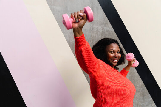 Happy plus size woman exercising with dumbbells near patterned wall