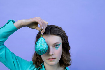 Teenage girl with make-up holding glittered pear in front of eye against lavender background