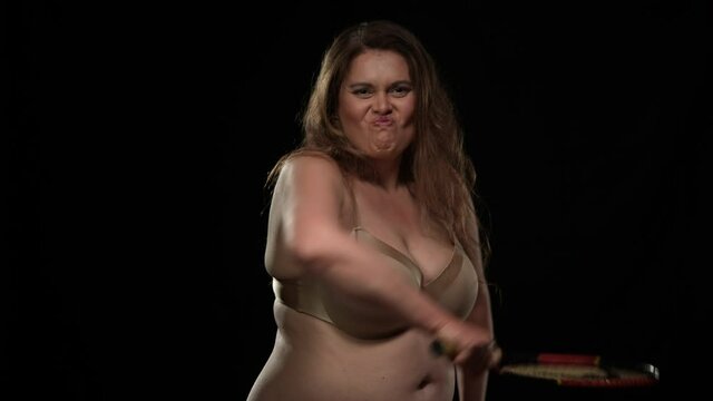 Angry furious plus size woman in bra looking at camera hitting with tennis racket. Portrait of aggressive irritated Caucasian obese lady posing at black background in underwear