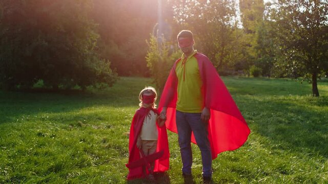 In the middle of the park in a sunny day posing in front of the camera cute small boy with his superhero daddy thy both wearing the red suits