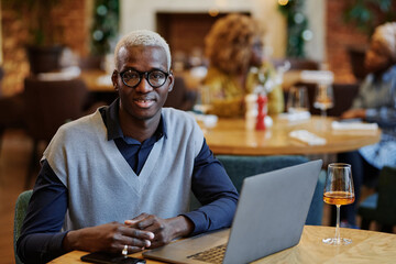 Portrait of African young businessman in eyeglasses smiling at camera while sitting at the table with laptop and glass of wine