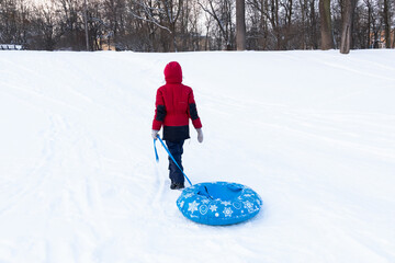 Boy in winter clothes walking with blue tubing among white snow, back view. Inflatable sleigh rides.