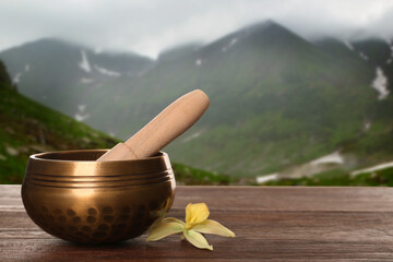 Golden singing bowl, mallet and flower on wooden table against mountain landscape. Space for text