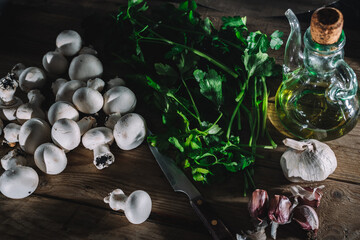 Piled up mushrooms and knife with garlic heads with loose cloves between parsley and canned oil with olive oil on rustic wood.