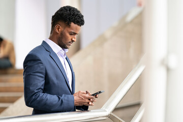 Businessman using mobile phone app texting outside of office in urban city with skyscrapers buildings in the background. Young black skin man holding smartphone for business work