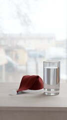 Santa's Christmas hat a glass of clear water on a wooden table in a bright room in front of the window