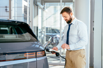 Beard caucasian man trying a new charging cable with a car charging station at the motor dealership. Concept of buying electric vehicle. Smart ecological living