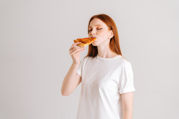 Studio portrait of happy attractive young woman with closed eyes appetite eating delicious pizza standing on white isolated background. Pretty redhead female eating tasty meal.