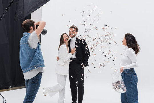 cheerful models in suits posing during photo session near assistant throwing confetti.