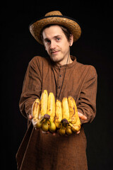 Young handsome tall slim white man with brown hair offering bunch of bananas with brown shirt and straw hat on black background