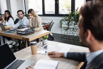 Obraz premium coworkers working at desk near laptop and businessman on blurred foreground.