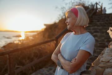 Side view of aged woman in sportswear, enjoying beauty of nature while looking at sea coast in evening sunset