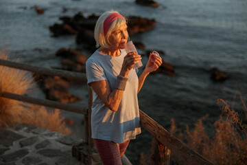Relaxed elderly lady wearing sports clothes drinking water and looking into distance, standing in autumn nature at sunset