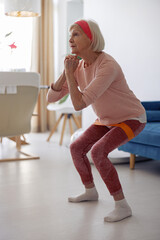 Active elderly lady working out while doing squats with elastic band on blurred interior of her...