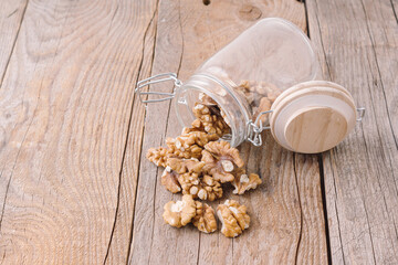 Fototapeta premium Peeled walnuts in glass jar on wooden background. Storage of nuts. Storage container at kitchen. Scattered walnuts. Space for text