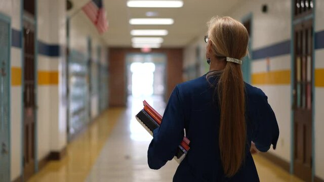 Slow motion rear view of teacher walking down a hallway in an empty school holding books showing emptiness.