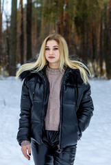 A young blonde girl in a black jacket with a fur hood stands in a winter forest, snow and a Christmas tree.