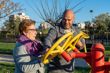 Male sports coach, helps an elderly woman to move the wheels in a bio-healthy park, at sunset. 