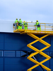 yellow self propelled scissor lift with workers against a blue sandwich panel wall background