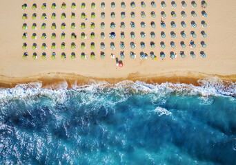 Top down view of beach with straw umbrellas. Umbrellas, sand and sea waves landscape. - obrazy, fototapety, plakaty